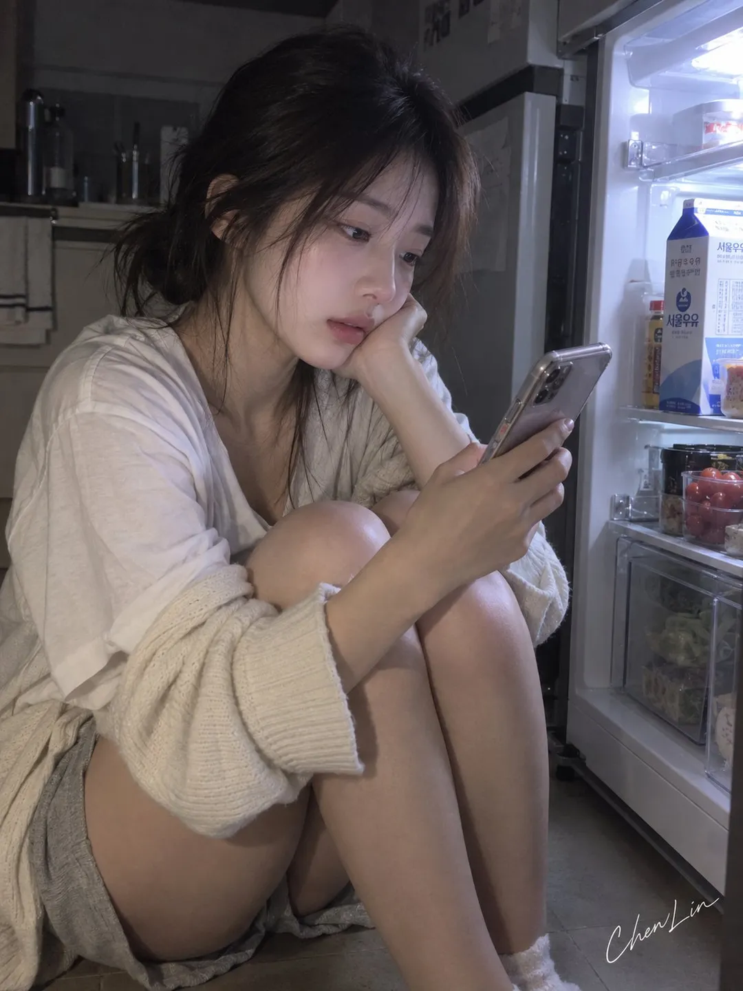 年轻女性穿着居家开衫和短裤蜷坐在深夜厨房冰箱旁查看手机｜通用 / 未注明 生成