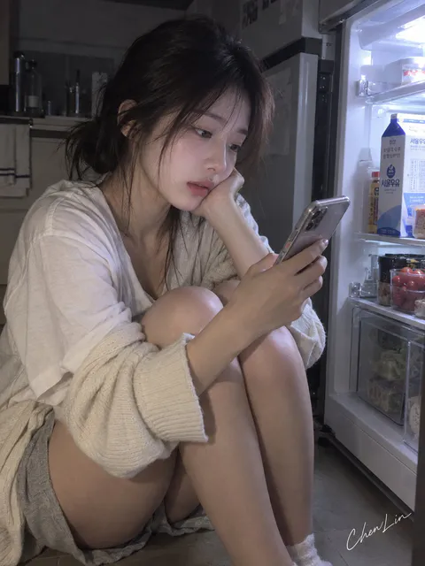 年轻女性穿着居家开衫和短裤蜷坐在深夜厨房冰箱旁查看手机｜通用 / 未注明 生成