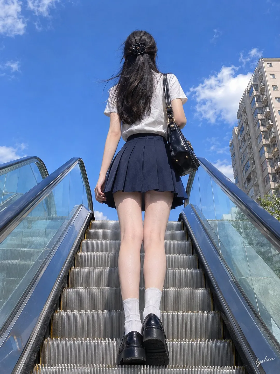 Low-angle shot from behind of a woman on an outdoor escalator wearing a white top and navy pleated skirt against a blue sky