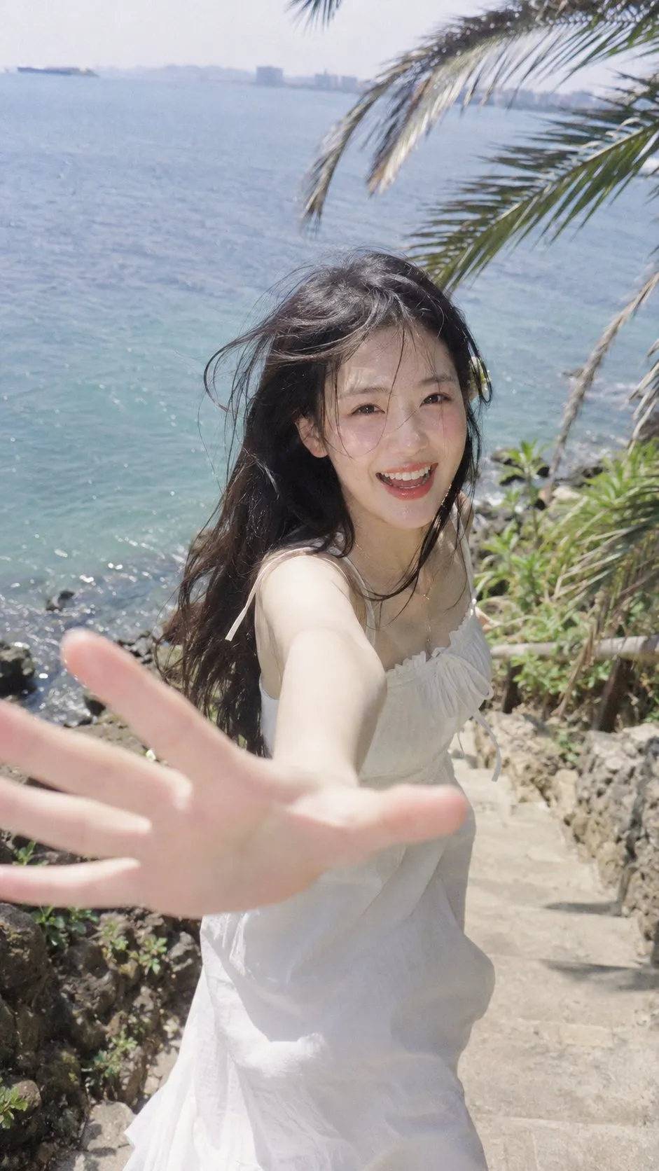海岛石阶上，身穿白色吊带裙的年轻女子笑着伸手探向镜头的俯拍抓拍。｜通用 / 未注明 生成