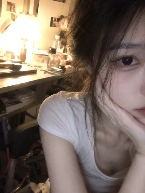 Close-up selfie of a young woman resting her chin on her hand in warm indoor desk lighting wearing a white t-shirt., generate