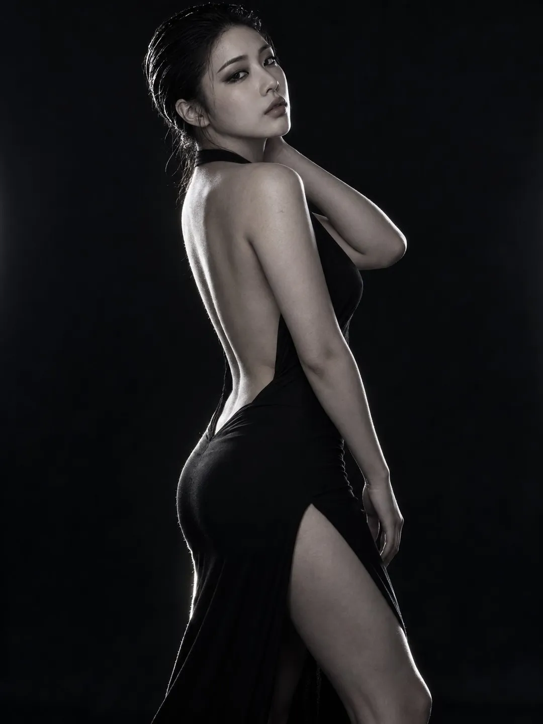A woman in a backless, high-slit black dress poses sideways in a dark studio with rim lighting highlighting her silhouette, g