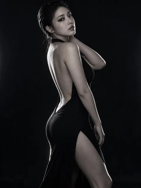 A woman in a backless, high-slit black dress poses sideways in a dark studio with rim lighting highlighting her silhouette, g